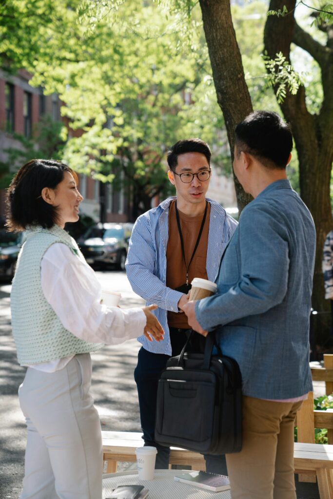 Three adults engaged in a friendly business meeting outdoors, shaking hands under green trees on a city street.